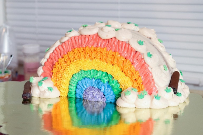 A half arch cake is decorated with icing in the colors of the rainbows. Icing clouds decorate the edges. on the "clouds" are cannabis shaped sprinkles and candy joints.