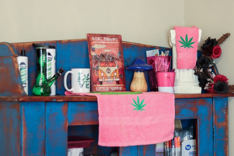 A shelf has various cannabis-related items. a bong, a cup with "Mama Mels Medibles" written on the side, a sign that reads "Stay Trippy Little Hippy" and some pink towels with marijuana leaves embroidered on the front.