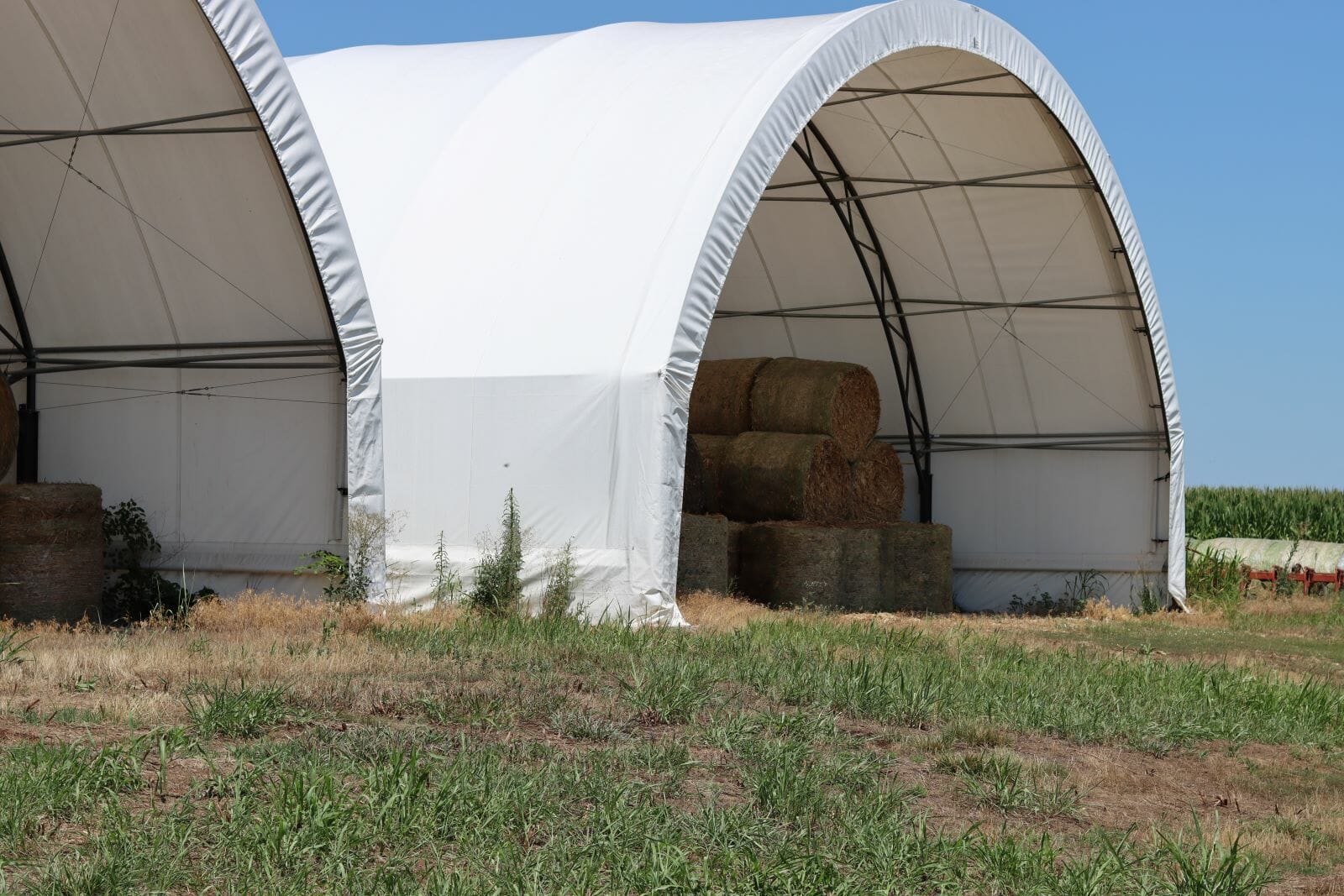 Two hoop barns covered in white tarp are filled with a small amount of round hay bales.