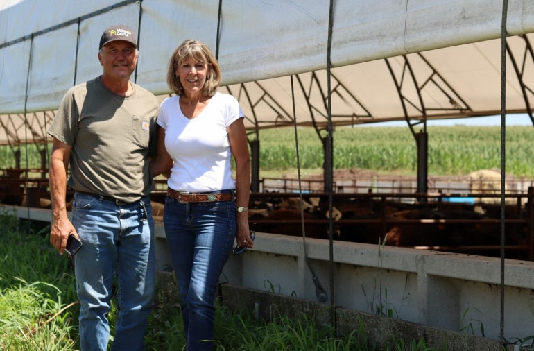 A man in a ball cap that reads "Mershon Cattle" stands next to a woman in a white shirt and jeans. They stand in front of a metal and white hoop barn full of young cattle.