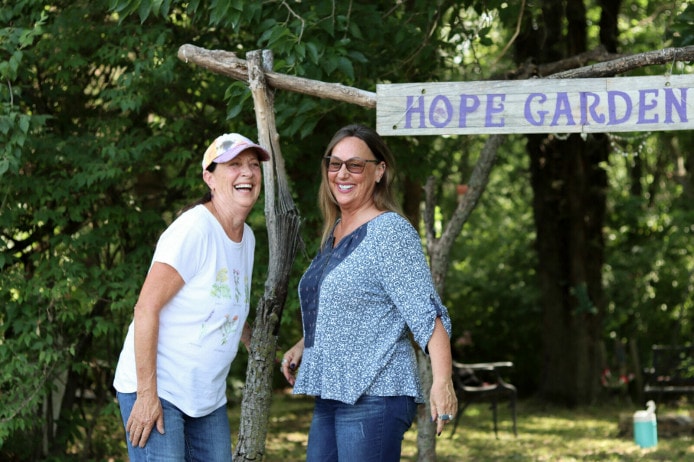 Two ladies laugh while standing in front of a natural arbor and a sign that reads "Hope Garden"