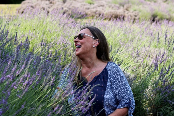 a woman with long hair, pink lipstick, sunglasses and a patterned shirt laughs while sitting among a field of lavender bushes.