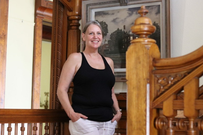 A woman in a black tank top stands next to an ornate wood staircase.