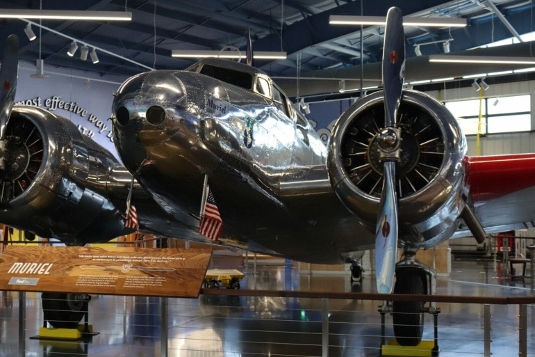 A big shiny silver plane with two prop engines. A sign in front of it reads "Muriel"