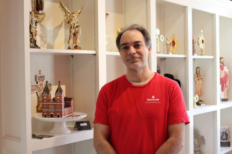 A man in a red shirt that reads "Benedictine" stands in front of shelves with religious figurines.