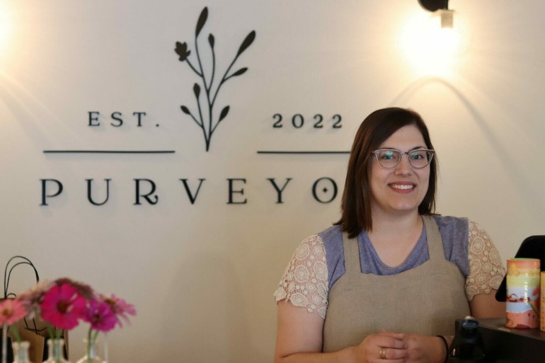 a woman with glasses and a linen apron stands in front of a sign that reads "Purveyor est. 2022)