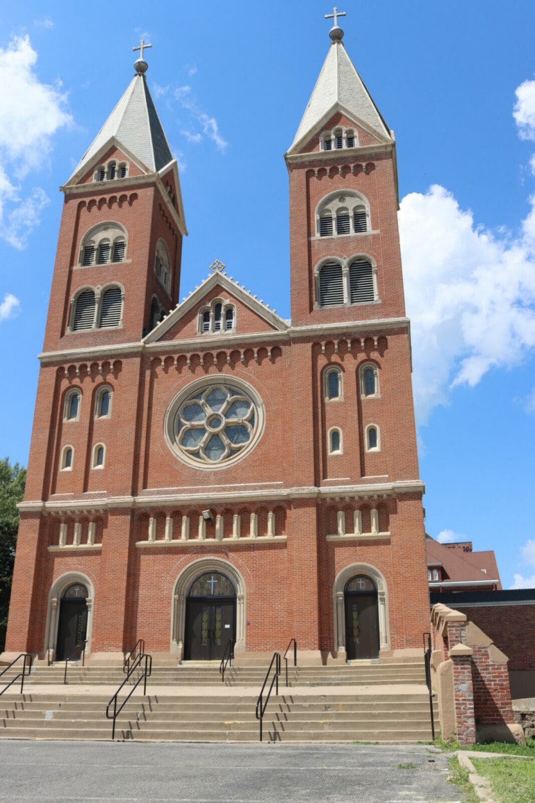 A red brick church has two towers and a rosary stained glass window in the center.