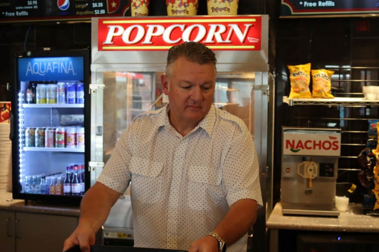 A man in a white button up stands in front of a movie concession stand. Behind him a big box reads "popcorn" and another reads "nachos"