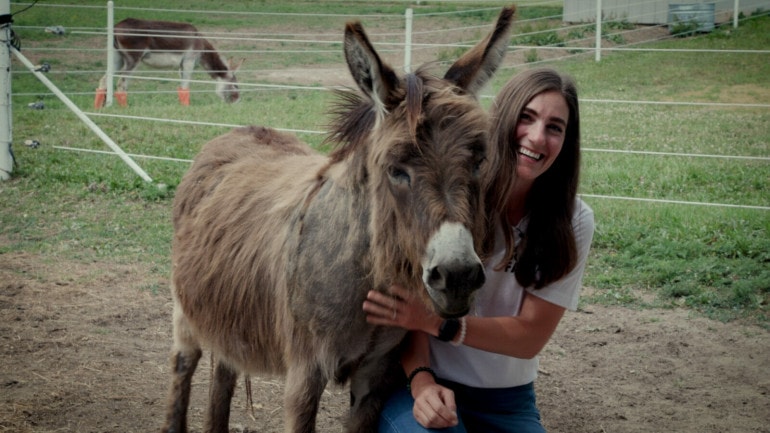A woman with dark hair crouches next to a short long haired donkey. She laughs while petting his beard.