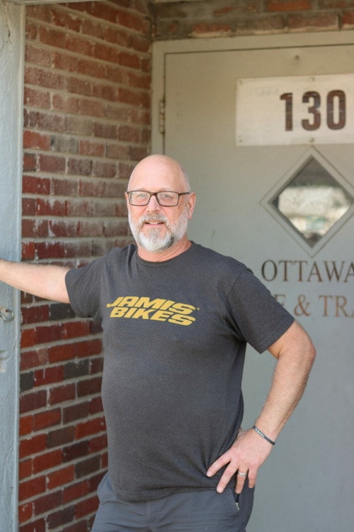 A man with a grey beard and bald head stands in front of a door that reads "Ottawa Bike and Trail." He has a grey shirt that reads "Jamis Bikes"