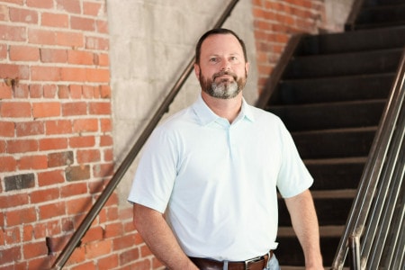 A man with a greying beard stands in front of a stairwell with exposed brick and concrete behind him.