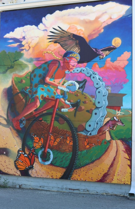 Colorful wall mural of a woman in a sunflower dress and a bike hat that reads "Kansas" on the up-turned bill. She is riding a bike along a gravel trail with monarch butterflies, haybales and other natural imagery.