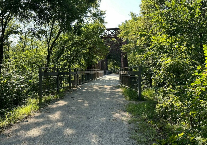 A gravel trail stretches to an oxidized steel bridge. Lush greenery hangs over the trail and partially obstructs the top section of the bridge.