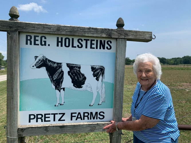 An older woman stands in a field next to a sign with a cow on it that reads "Reg. Holsteins Pretz Farms"
