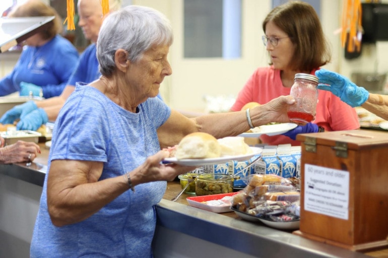 At the end of a food line, an aging woman holds her plate an accepts a jar of homemade jam from someone behind the counter.