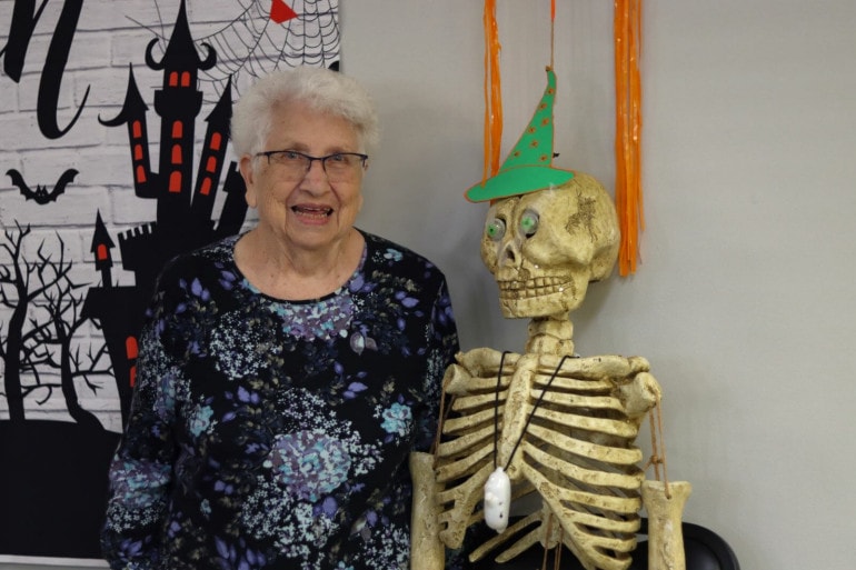 An older woman with glasses and a floral shirt stands next to a skeleton and other Halloween decorations.