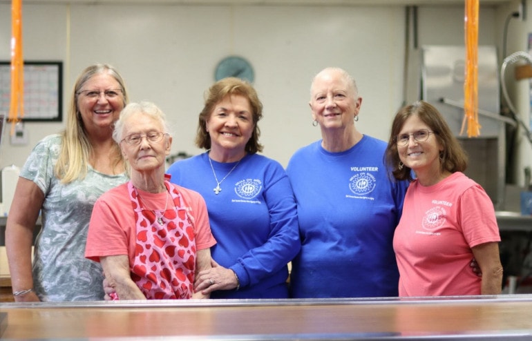 A group of women smile from behind a kitchen. Many of them wear shirts that read, "Meals on Wheels Volunteer"