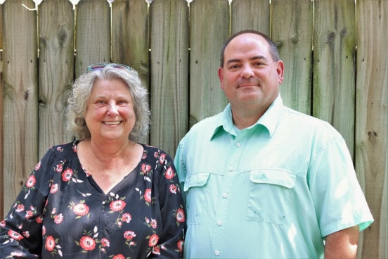 an older woman with gray hair and a floral shirt stands next to a middle aged man with a button up shirt on.
