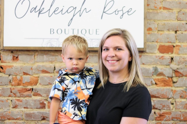 A young woman with blonde hair holds a toddler who scowls at the camera. Behind her is a sign that reads Oakleigh Rose Boutique.
