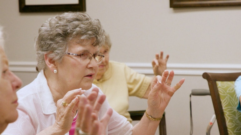 an older woman with glasses holds up her hands and touches her forefinger to her thumb.