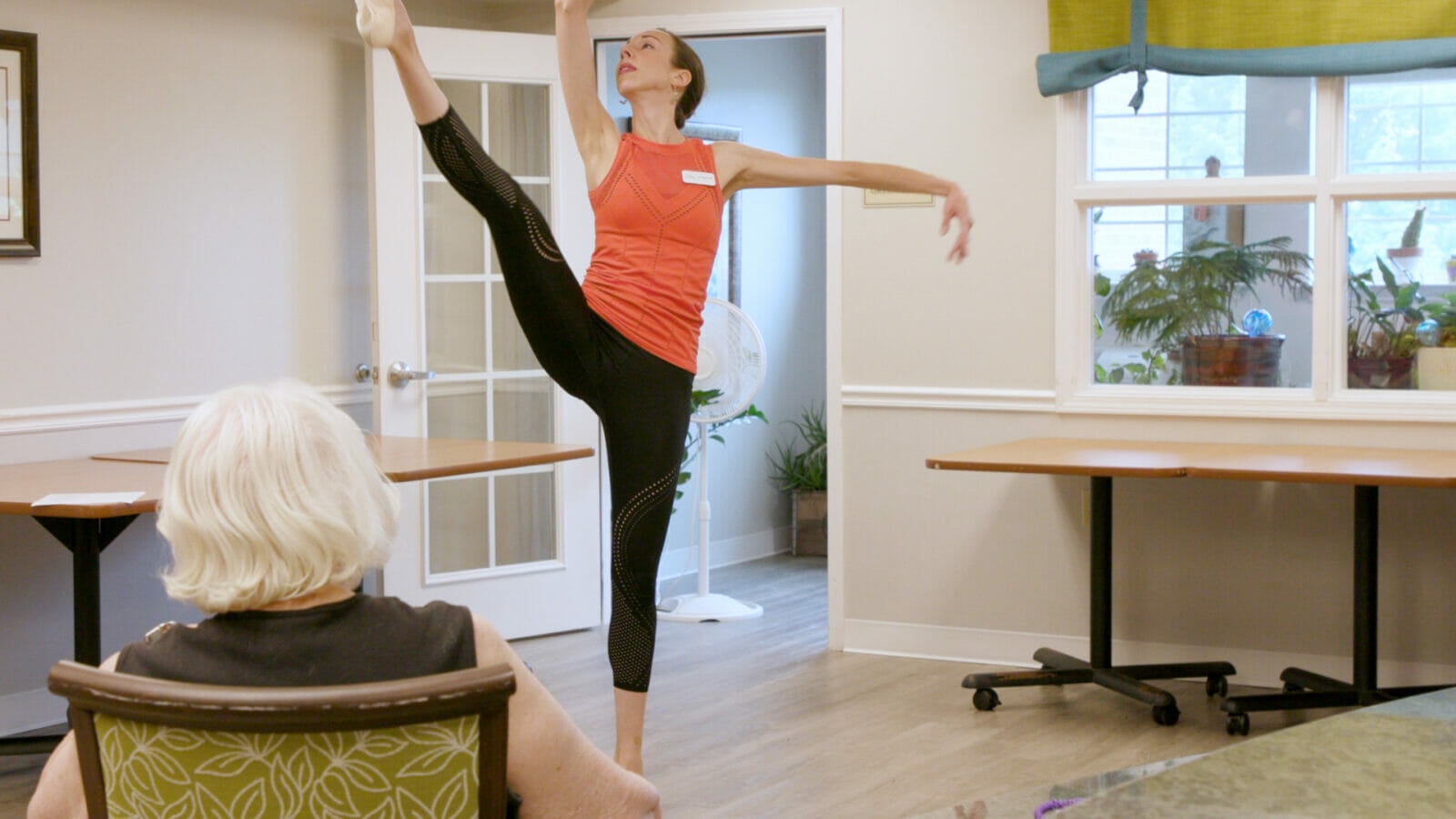 A woman in an orange shirt and black leggings kicks her leg up high in the air in a ballet move at the front of a room.