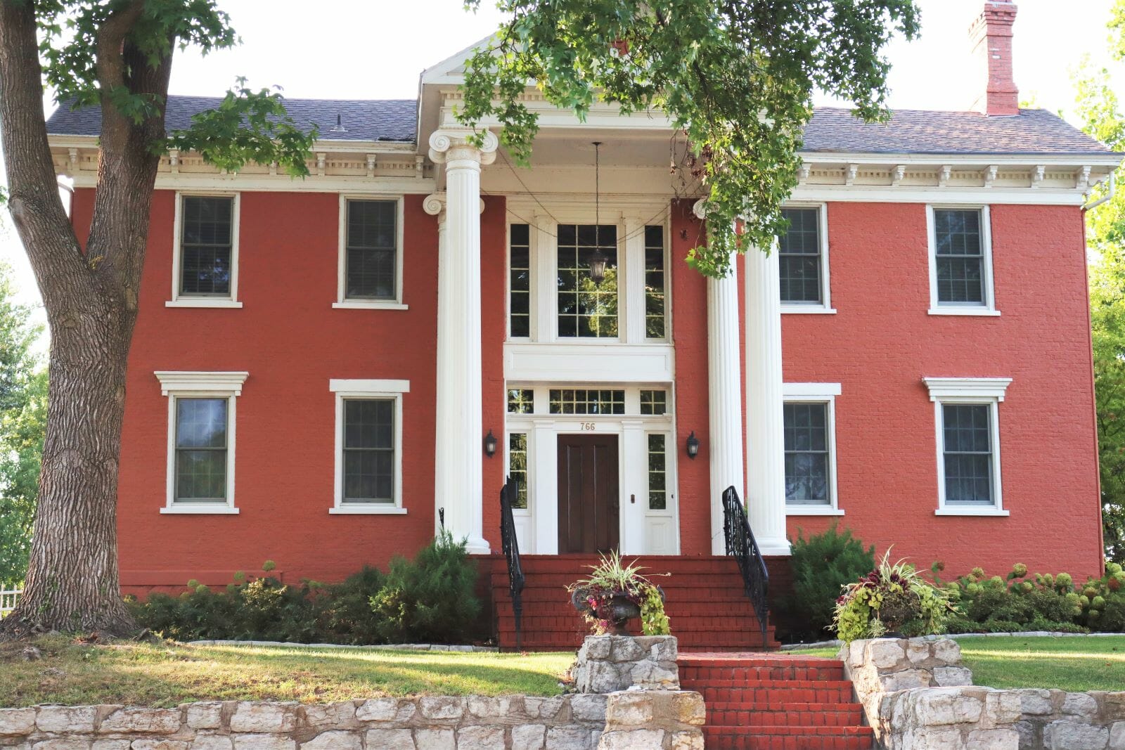 a large painted red brick home with white columns, windows and detailing. The home features ornate iron railing on the stairs leading up to the front door.