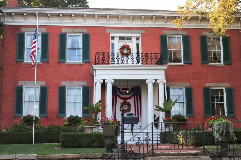 Another Greek revival style house with painted red brick, white columns and green window shutters. a balcony above the front door has an ornate iron fence.