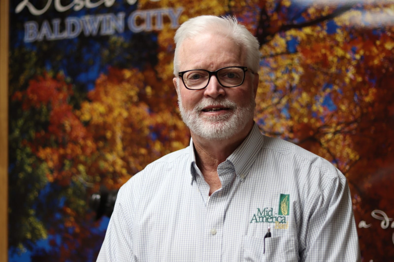 A man with glasses stands in front of a poster of an orange and red maple tree. His shirt reads "Mid America Bank"