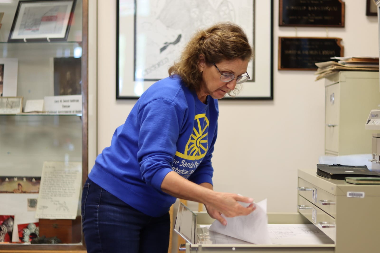 A woman in glasses peers over a pull out drawer and flips through a stack of maps.