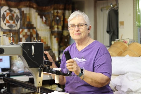 an older woman in a purple shirt stands at a long arm quilt machine