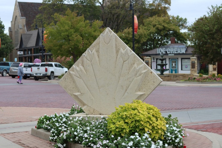 A stone statue with a carving of a maple leaf on it stands on the corner of a block. Behind it are stone pillars, brick streets and downtown buildings.