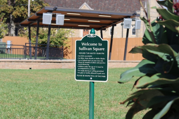 A green sign reads "Welcome to Sullivan Square" behind it is a covered stage in a grass yard.