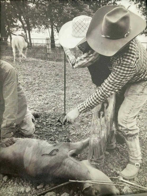 An man stands behind a young boy and points towards a calf, which lies on the ground. The young man is holding a branding iron and the calf is branded with the letter "J". Both are wearing cowboy attire.