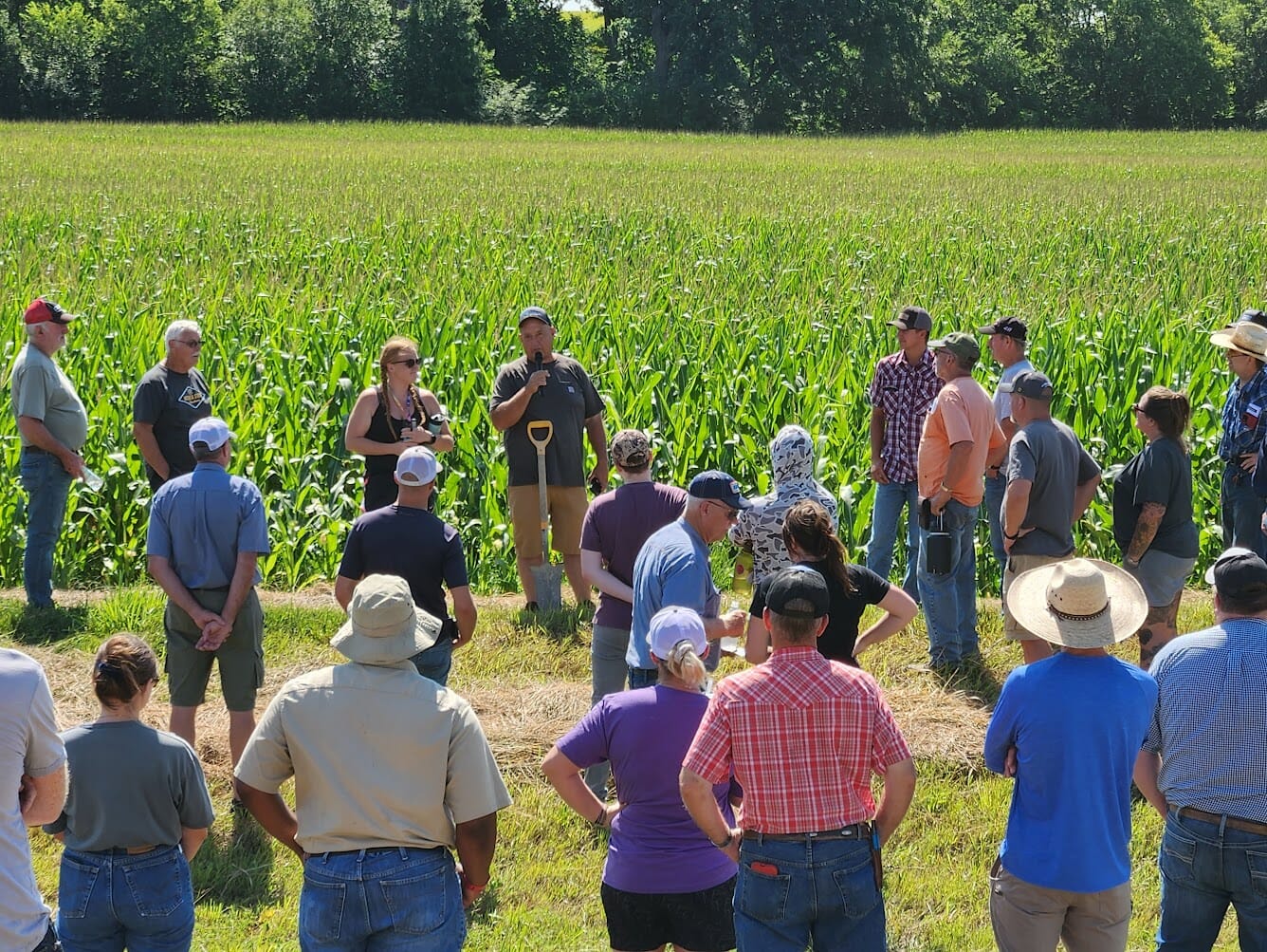 a crowd of about 20 people stand in front of a corn field. A man at the front of the crowd has a shovel and speaks into a microphone.