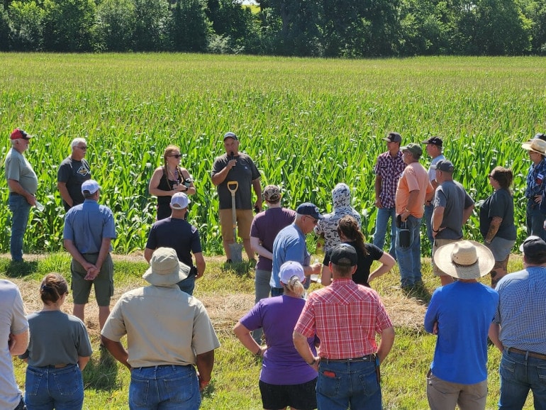 a crowd of about 20 people stand in front of a corn field. A man at the front of the crowd has a shovel and speaks into a microphone.