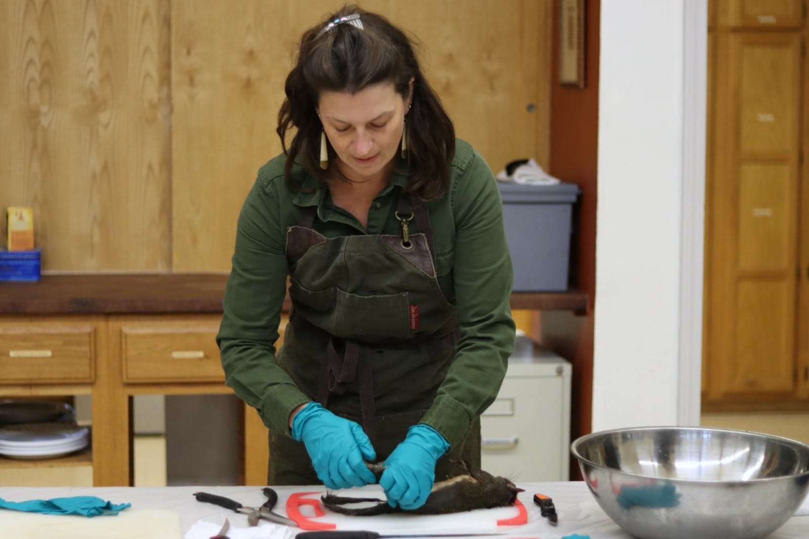 A woman in a green top and green apron feels for the joint on a squirrels leg that lies (dead) on a cutting board.