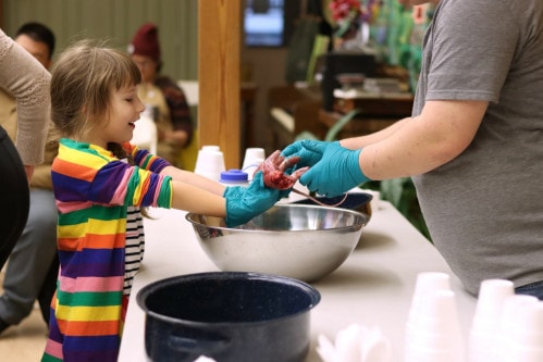 A young girl in a colorful striped shirt holds up a skinned and gutted squirrel from a silver bowl.