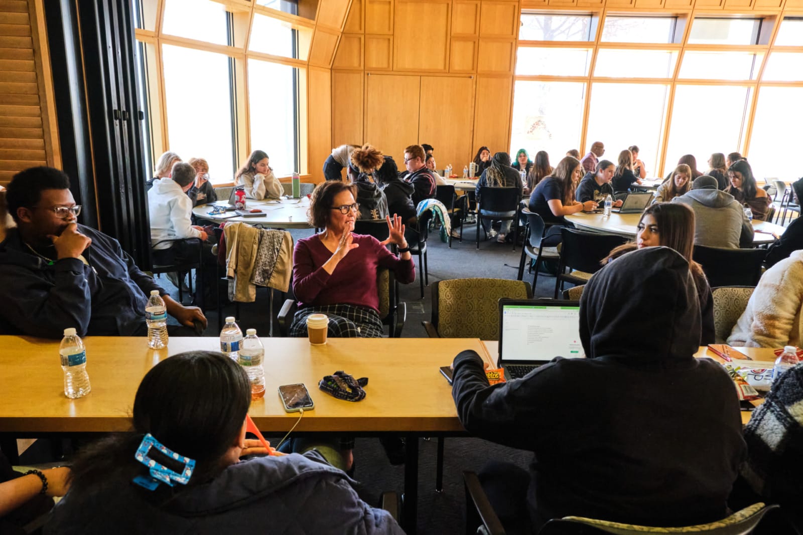 A wide shot of students sat around a large group of tables. Each table is engaged in conversation and research.