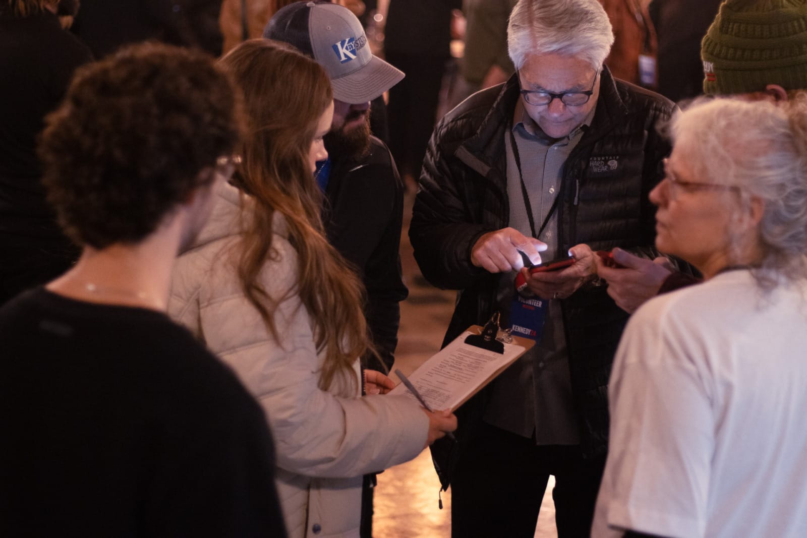 A group of people crowd around a clipboard. One woman holds the clipboard with a pen in her hand as well.