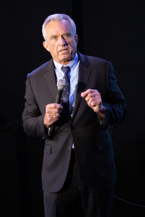 A man in a black suit with blue shirt and tie. He holds a microphone in one hand.
