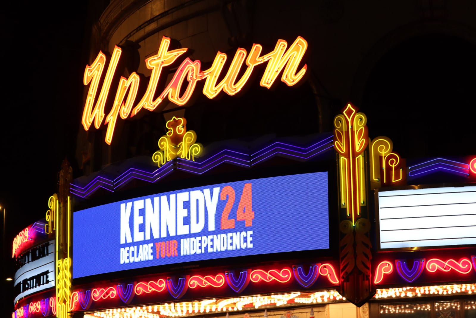 Neon lights on a building read "Uptown" an electronic marquee reads "Kennedy 24 Declare your Independence"