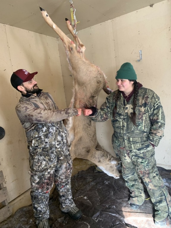 Two people with camo coveralls on fist bump and smile. behind them hangs a dead deer.