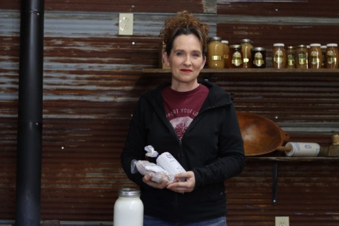 A woman in a black jacket and jeans holds a package of meat with a jar of milk next to her. Behind her is a tin wall with jars of honey.
