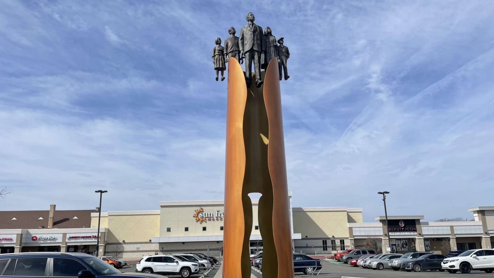 A tall bronze memorial with figures at the top in front of Sun Fresh market.
