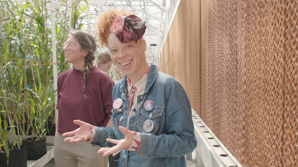 A woman with red hair and a scarf smiles and gestures with her hands. Behind her are rows of tall sorghum plants and a woman with a maroon tshirt.