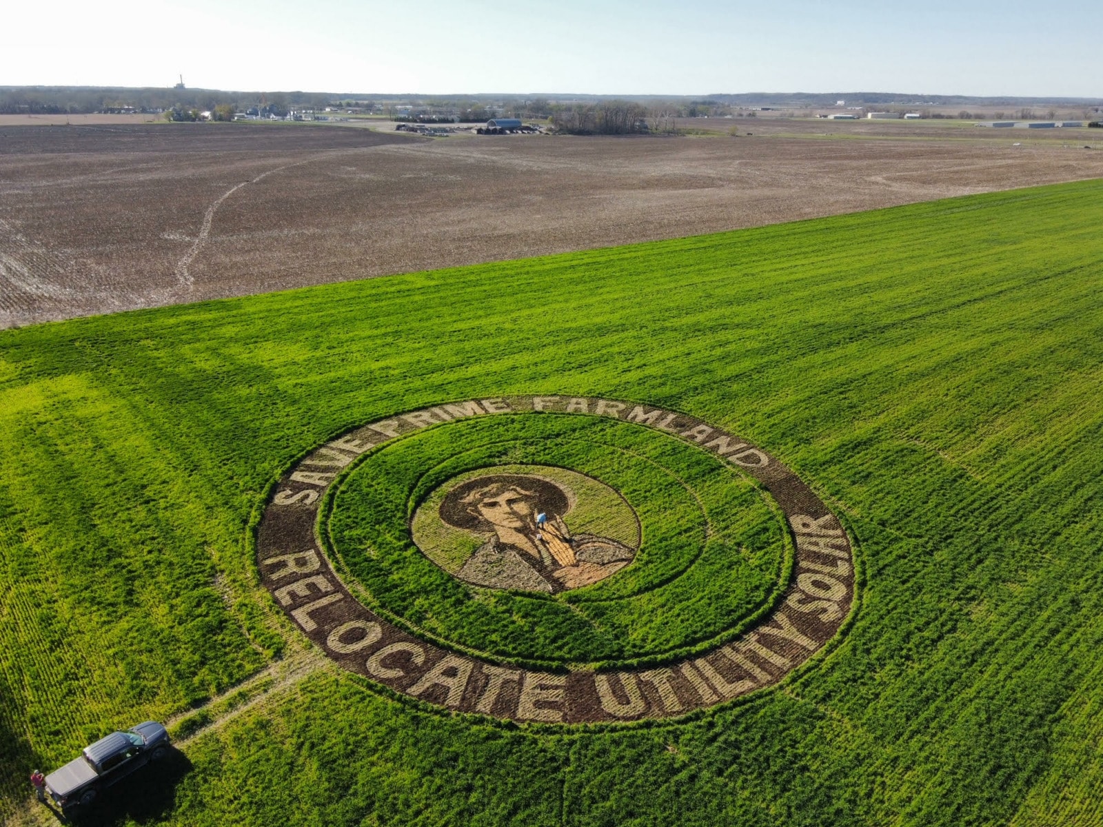 A green field. In the middle of the field is an art piece made of dirt and grass that depicts a young farmer. Around the outside of the circle it reads "Save Prime Soil" "Relocate Utility Soil"