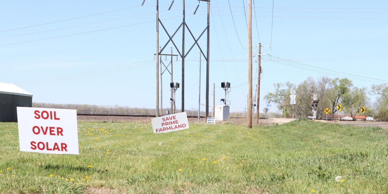 Three white signs in the grass sit in front of railroad tracks and powerlines. The signs read, "Soil over solar" "save prime farmland" and "Relocate Utility Solar"