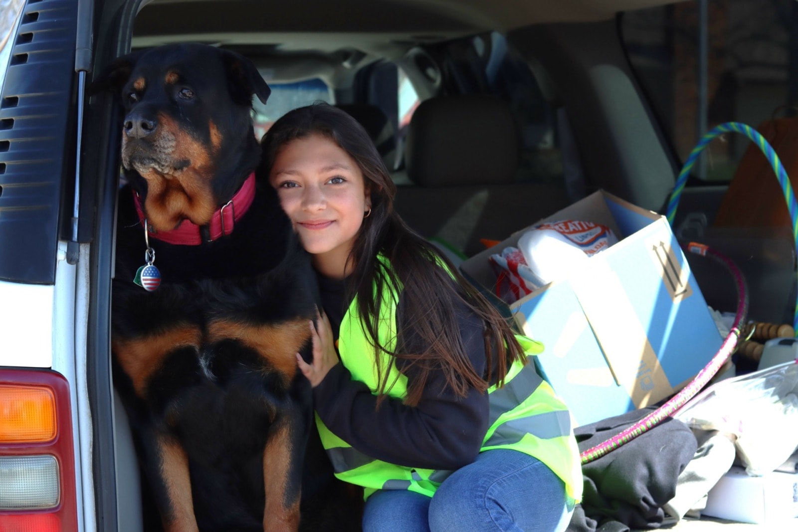 A young girl sits in the back of an SUV and hugs her large black and brown dog.