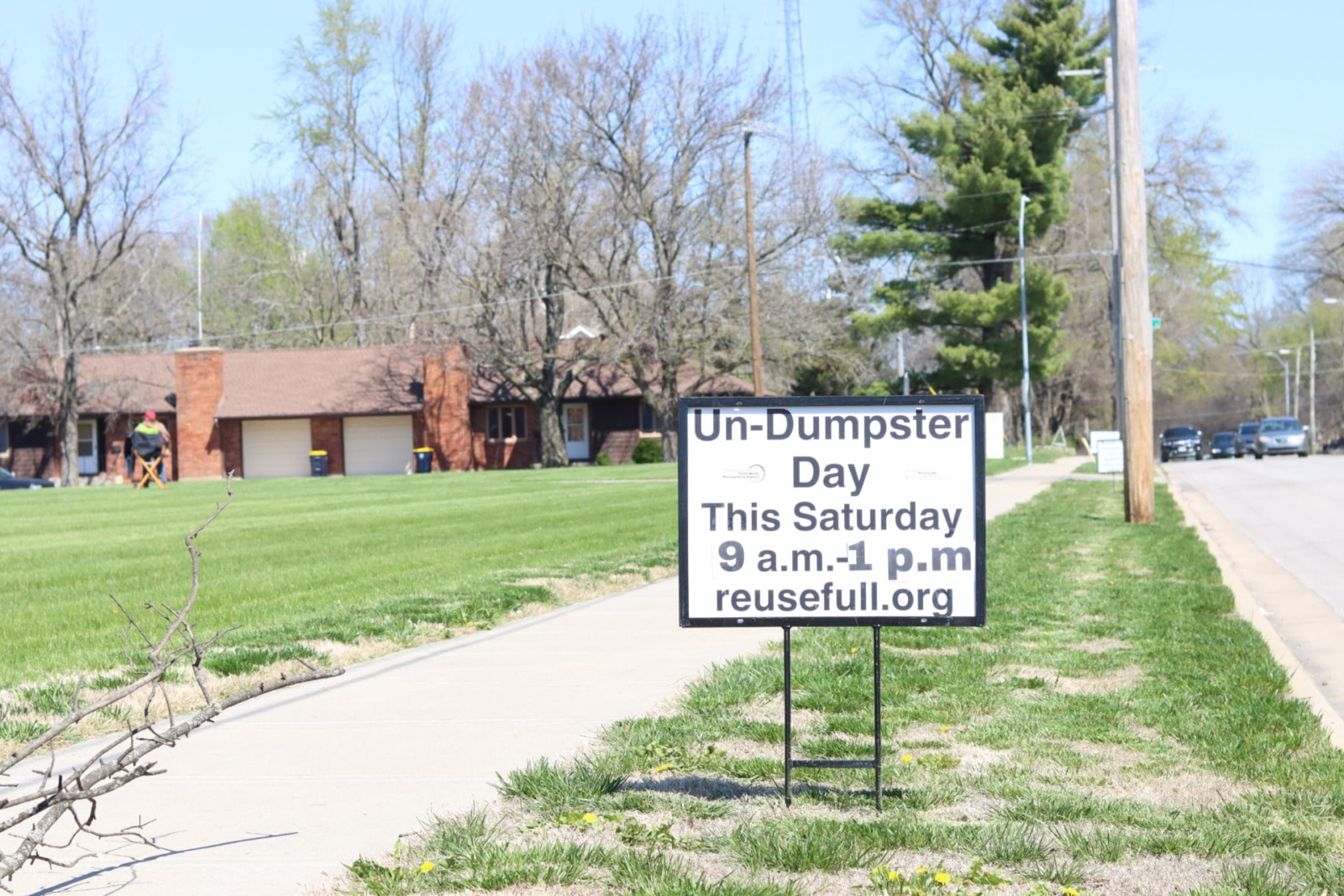 A sign along a sidewalk reads "Un-Dumpster Day This Saturday 9 a.m. - 1 p.m. reusefull.org"