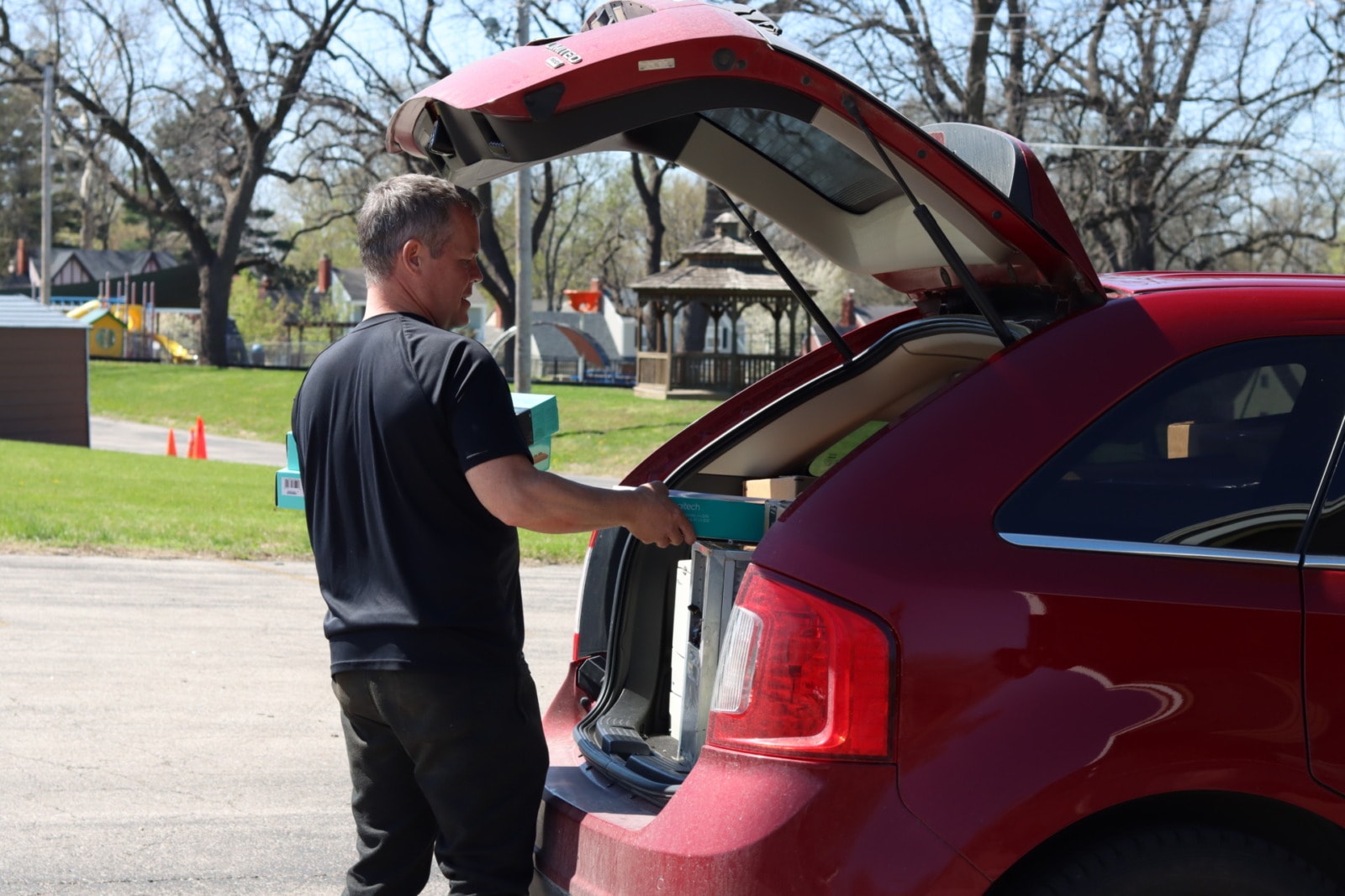 A man in a black shirt unloads small boxes from the back of a red car.